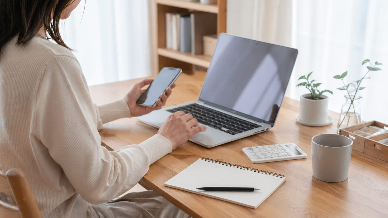 自宅デスクでノートパソコンとスマホを使って保険を調べる女性